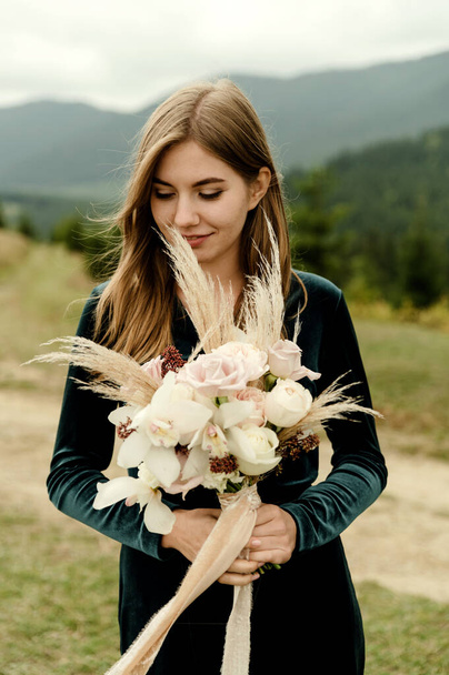 portrait of the bride. A young girl in a dark green wedding dress with a bouquet of flowers and greenery in her hands against the backdrop of mountains and forests at sunset - Photo, Image