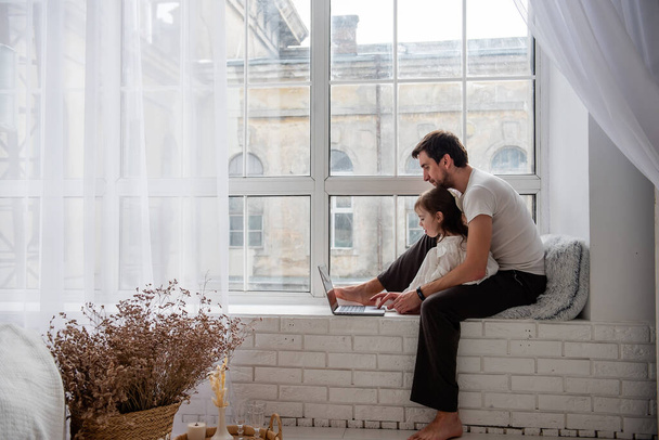 Father and daughter in pajamas are playing on laptop, sitting on wooden, white windowsill near the window. Young man teaches little girl how to work on computer. Technological generation of the family - Photo, Image