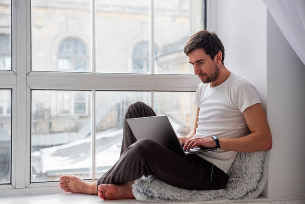 A young handsome man sits on a wooden windowsill near the window in a lotus position, works on a gray laptop. Remote work freelancer. The programmer, hacker is typing on a computer at home in pajamas - Foto, afbeelding