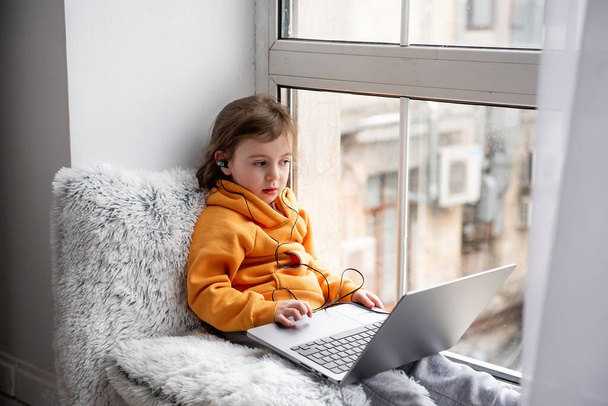 Mała dziewczynka siedzi na szerokim parapecie, gra w gry na laptopie, słucha muzyki na słuchawkach. Dziecko w pomarańczowej bluzie, szare spodnie przy oknie domu. Kształcenie na odległość dla przedszkolaków - Zdjęcie, obraz