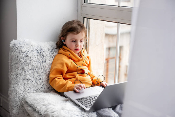 Little toddler girl sits on wide windowsill, plays games on laptop, listens to music on headphones. Child in an orange hoodie, gray pants at the window of the house. Distance learning for preschoolers - Zdjęcie, obraz