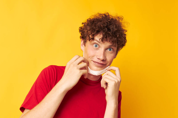 guy with red curly hair wearing a red t-shirt medical mask on the face posing yellow background unaltered - Foto, Imagen