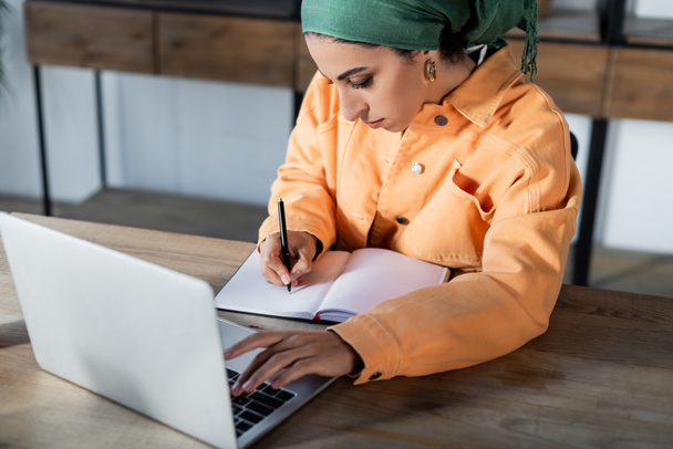 muslim γυναίκα που γράφει σε κενό σημειωματάριο, ενώ χρησιμοποιώντας το φορητό υπολογιστή στο σπίτι - Φωτογραφία, εικόνα