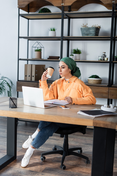 muslim woman holding paper cup while sitting near laptop at home - Photo, Image