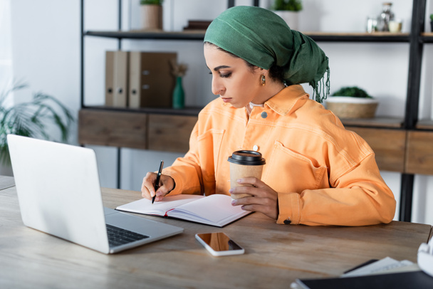 mujer musulmana con café para ir escribiendo en un cuaderno vacío cerca de la computadora portátil y el teléfono móvil - Foto, Imagen
