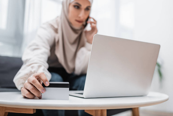 foco seletivo do cartão de crédito na mão de mulher muçulmana falando no celular perto do laptop - Foto, Imagem