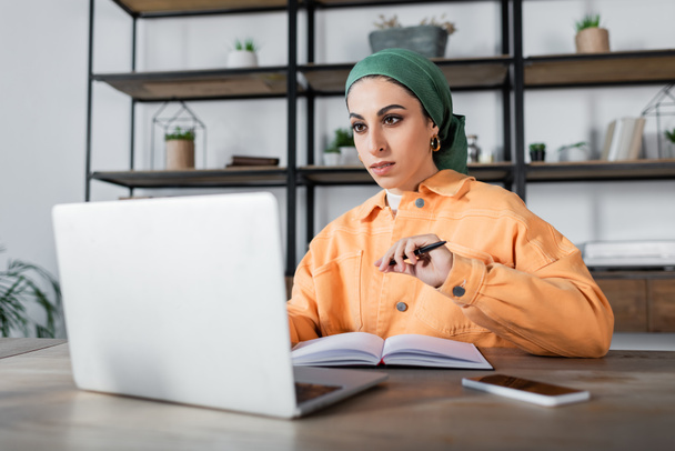 muslim woman in headkerchief and orange jacket sitting with pen near blurred notebook - Fotoğraf, Görsel