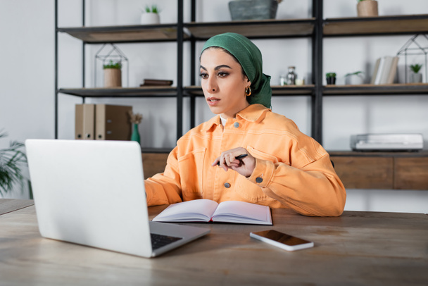 arabian woman in headkerchief looking at laptop near empty notebook on desk - Photo, Image