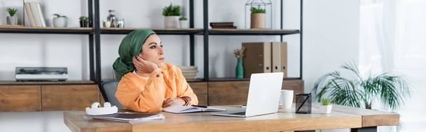 mulher muçulmana olhando para longe enquanto sentado perto de laptop em casa, banner - Foto, Imagem