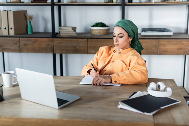 Evdeki dizüstü bilgisayarın yanında çalışırken mendilli müslüman bir kadın deftere yazı yazıyor. - Fotoğraf, Görsel