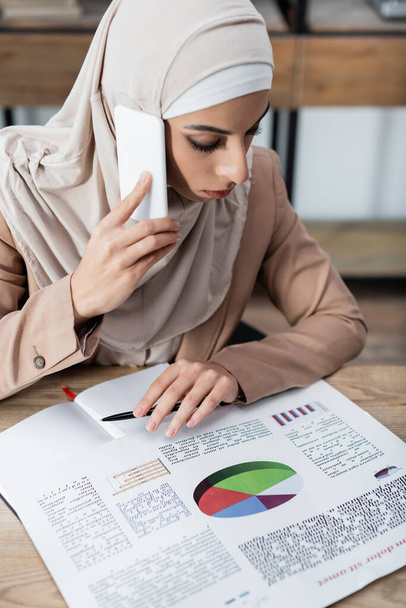 arabian businesswoman in hijab talking on smartphone while working with infographics at home - Photo, Image