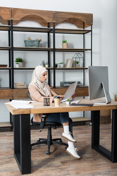Müslüman kadın evdeki bilgisayarın yanında otururken belgeye bakıyor - Fotoğraf, Görsel
