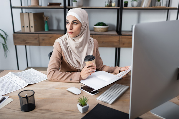 mujer musulmana mirando hacia otro lado y sosteniendo café para ir mientras está sentado en la oficina en casa - Foto, imagen
