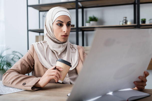 Araberin im Hijab mit Pappbecher und verschwommenem Dokument bei der Arbeit zu Hause - Foto, Bild