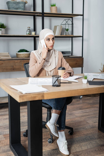 muslim woman talking on smartphone while sitting near coffee to go on work desk at home - 写真・画像
