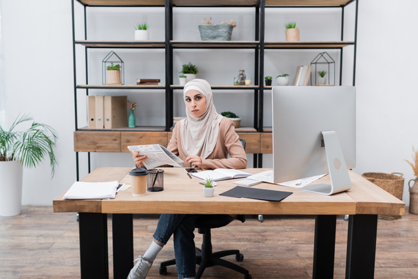 muslim woman holding newspaper and looking at camera near computer on work desk at home - Photo, Image