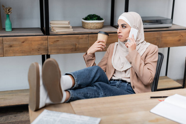 muslim woman talking on smartphone while sitting with legs on desk at home - Photo, Image