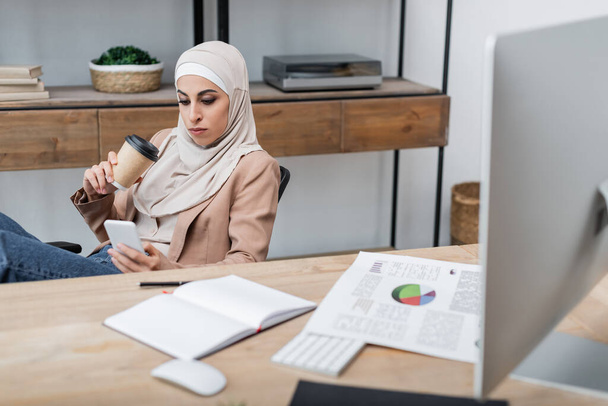 muslim woman with coffee to go and smartphone sitting near blank notebook and charts on desk - Foto, imagen