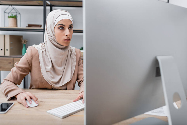mulher muçulmana trabalhando perto de monitor de computador borrado e smartphone com tela em branco em casa - Foto, Imagem