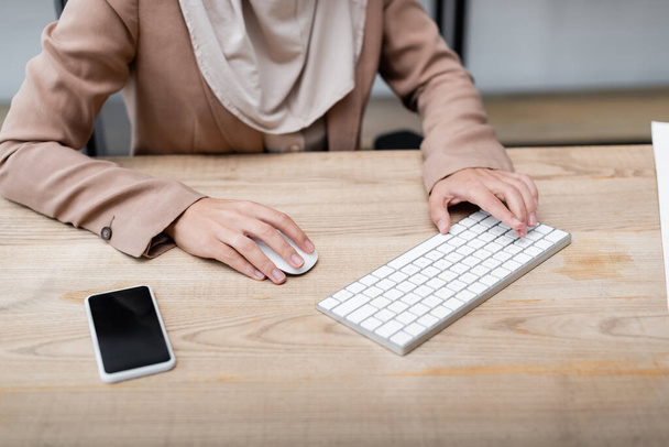 Ausgeschnittene Ansicht einer Frau, die Tastatur und Computermaus benutzt, während sie in der Nähe des Smartphones mit leerem Bildschirm arbeitet - Foto, Bild