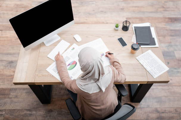 top view of arabian woman in hijab working with charts near monitor with blank screen - Zdjęcie, obraz