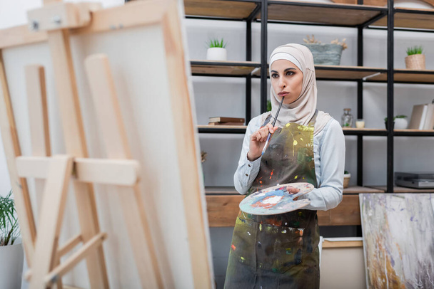 mujer árabe pensativa con paleta y pincel pensando cerca del caballete en casa - Foto, imagen