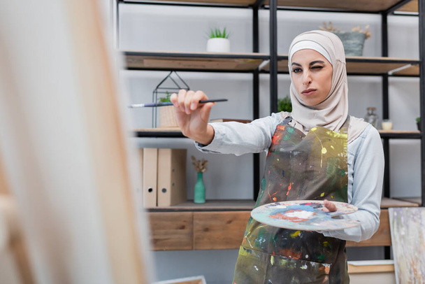mujer musulmana con paleta y pincel cerca de lienzo borroso en casa - Foto, Imagen