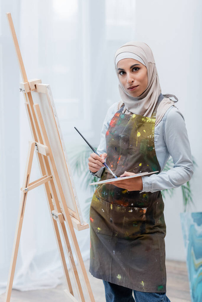 arabska kobieta z paletą i pędzlem patrząca w kamerę w pobliżu sztalugi - Zdjęcie, obraz