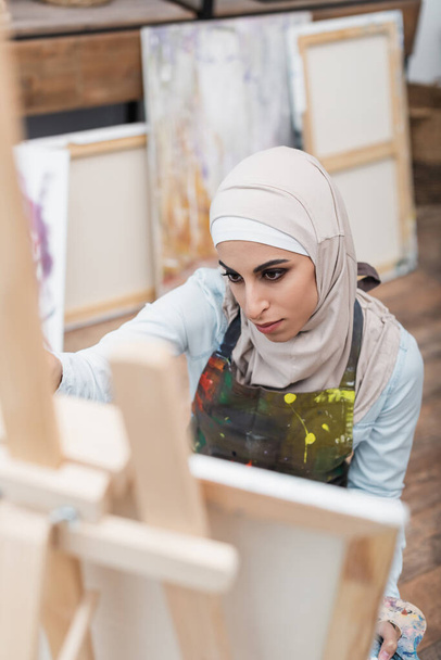 vista ad alto angolo della donna araba in hijab disegno su cavalletto sfocato a casa - Foto, immagini