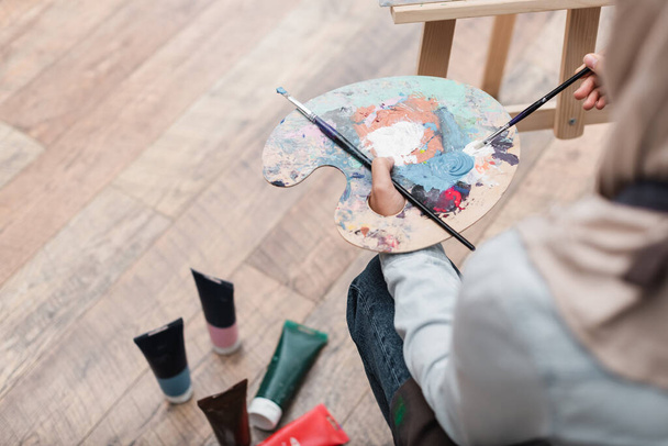 high angle view of blurred woman with palette and paintbrushes near paint tubes on floor - Foto, Imagen