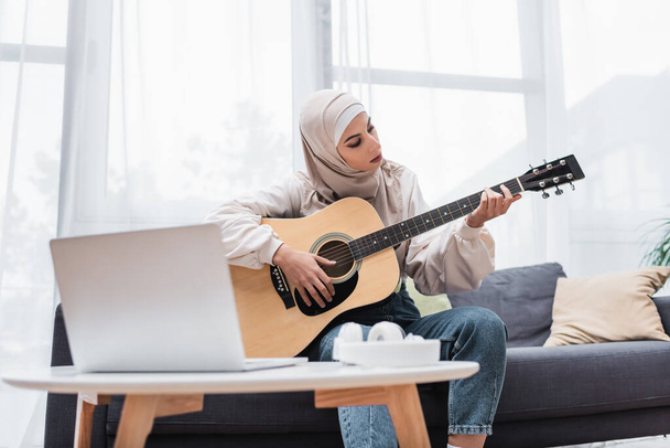 Muslimische Frau spielt Gitarre, während sie auf dem Sofa sitzt, während der Videostunde auf dem Laptop - Foto, Bild