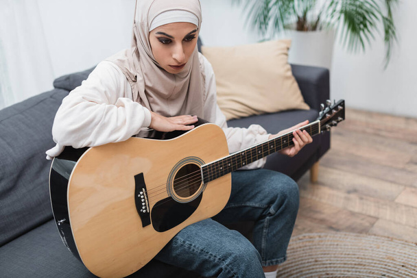 Muslimische Frau mit Akustikgitarre sitzt zu Hause auf Couch - Foto, Bild
