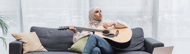 arabian woman looking away while sitting on couch and playing guitar, banner - Foto, Bild