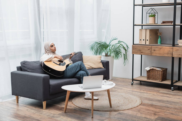 Άραβα γυναίκα με μαντίλα παίζει κιθάρα στον καναπέ κοντά στο λάπτοπ - Φωτογραφία, εικόνα