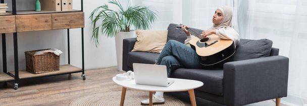 mladá muslimka hraje na kytaru, zatímco sedí na gauči u notebooku v obývacím pokoji, banner - Fotografie, Obrázek