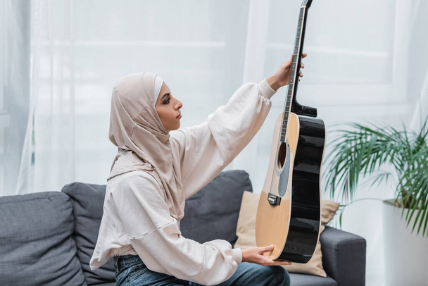 side view of muslim woman looking at guitar while sitting on couch at home - Foto, immagini