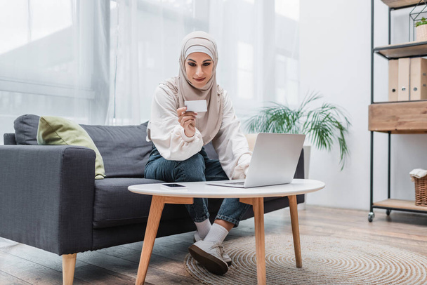smiling arabian woman looking at credit card while sitting on sofa near laptop - Photo, Image