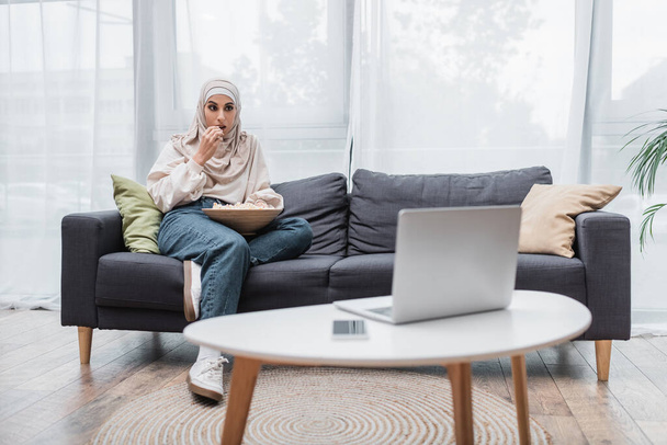 Мусульманка сидит на диване и ест попкорн во время просмотра фильма на ноутбуке - Фото, изображение