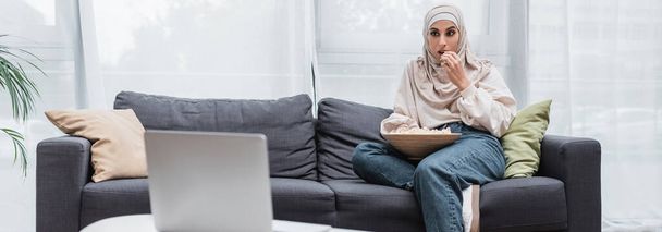 mujer árabe en hijab comiendo palomitas de maíz y viendo la película en el ordenador portátil en casa, pancarta - Foto, imagen