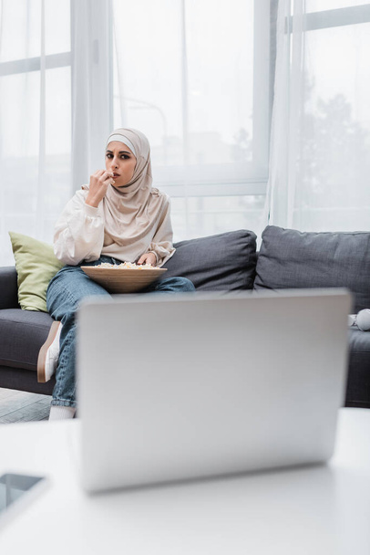 concentrated muslim woman eating popcorn while watching movie on blurred laptop - Foto, afbeelding