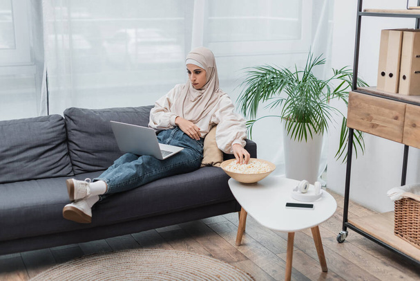 Арабианка смотрит кино на ноутбуке рядом с попкорном и смартфоном на кофейном столике - Фото, изображение