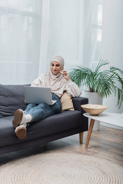 веселая арабская женщина в хиджабе ест попкорн и смотрит фильм на ноутбуке на диване - Фото, изображение