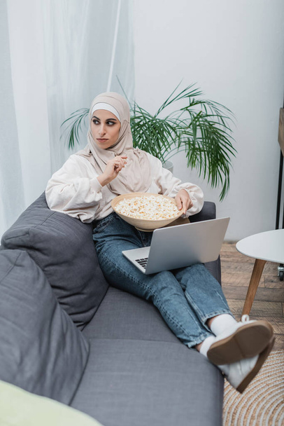 mujer musulmana mirando hacia otro lado mientras está sentado en el sofá con palomitas de maíz y portátil - Foto, Imagen