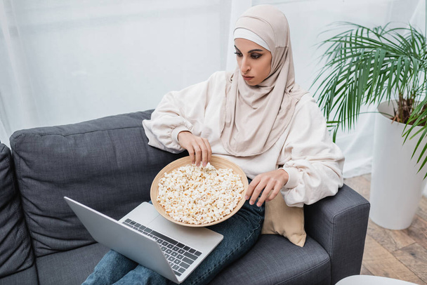 arabian woman in hijab watching movie on laptop while sitting on sofa with bowl of popcorn - Zdjęcie, obraz