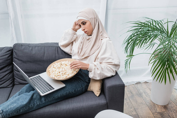 σκεπτική αραβική γυναίκα με μπολ ποπ κορν βλέποντας ταινία στο laptop κοντά στο πράσινο potted παλάμη - Φωτογραφία, εικόνα