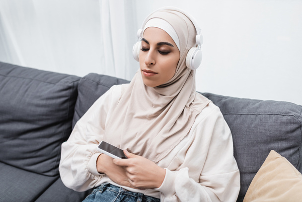 arabian woman listening music in headphones while sitting on couch with closed eyes - Fotografie, Obrázek