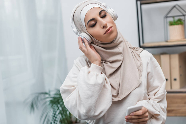młoda muzułmanka w hidżabie i słuchawki słuchające muzyki z zamkniętymi oczami - Zdjęcie, obraz