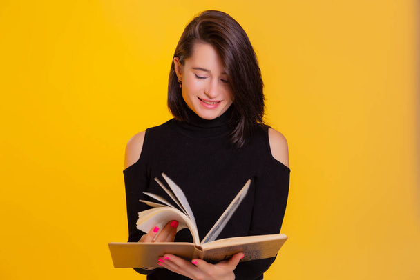 Hübsche Frau mit einem Buch in der Hand, das in die Kamera lächelt. Kluge und aufgeregte Dame mit offenem Buch. Studioaufnahme isoliert auf gelbem Hintergrund - Foto, Bild
