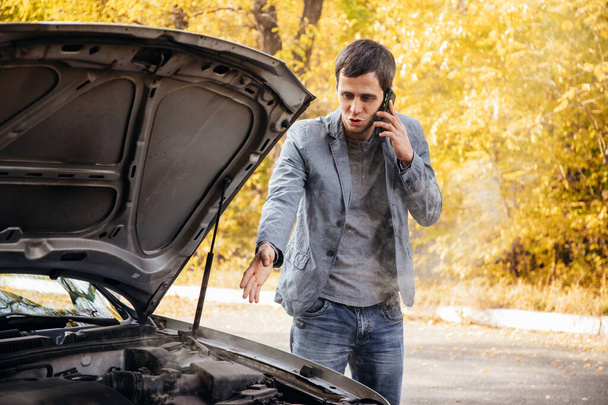 A man looks under the open hood of a car. The car broke down on the road. The engine is smoking - Foto, Imagen