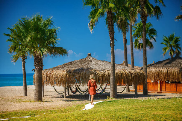 Sahilde bungalovlara doğru yürüyen plaj elbiseli sarışın bir kadın. - Fotoğraf, Görsel